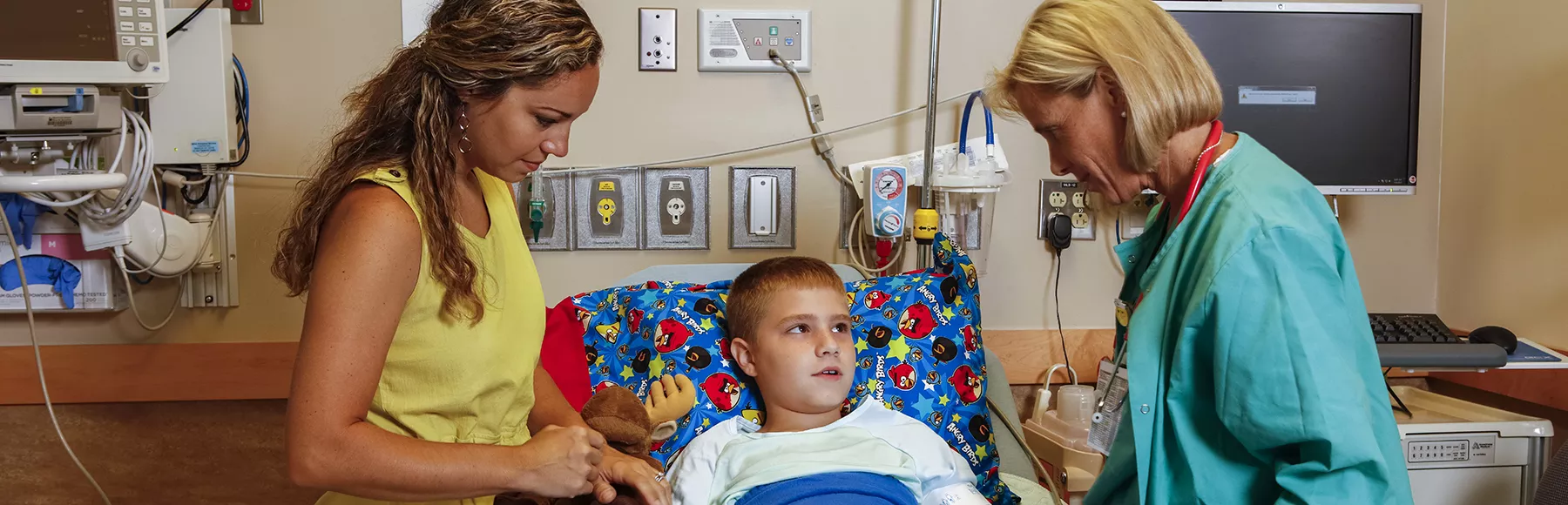 photo: mom standing beside hospital bed, boy on bed looking at nurse standing on other side of bed